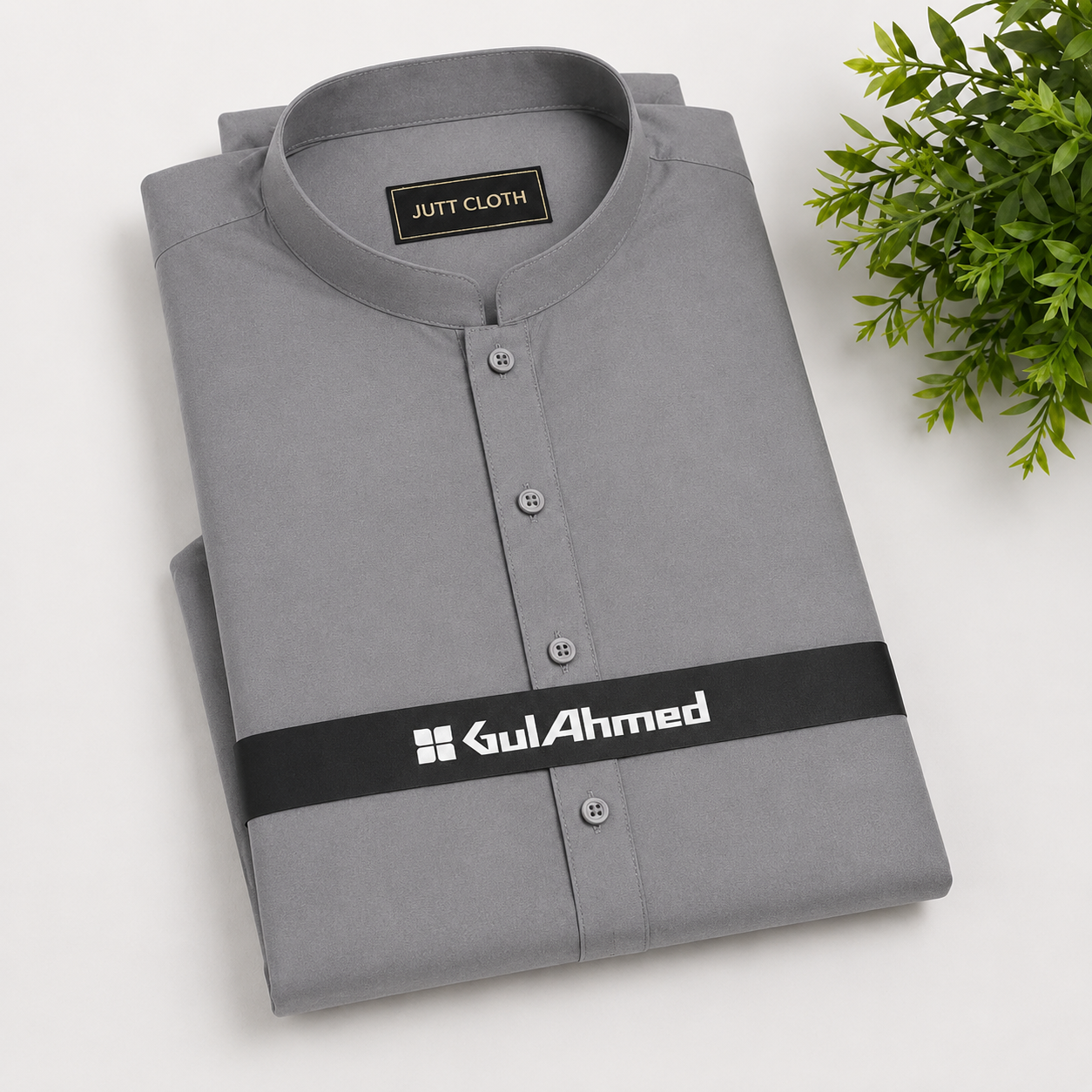 A folded gray long-sleeve shirt with a mandarin collar and silver buttons, featuring a black waist strap with the brand name 'GulAhmed' in white. The shirt is displayed on a light gray background with a green plant in the top right corner.