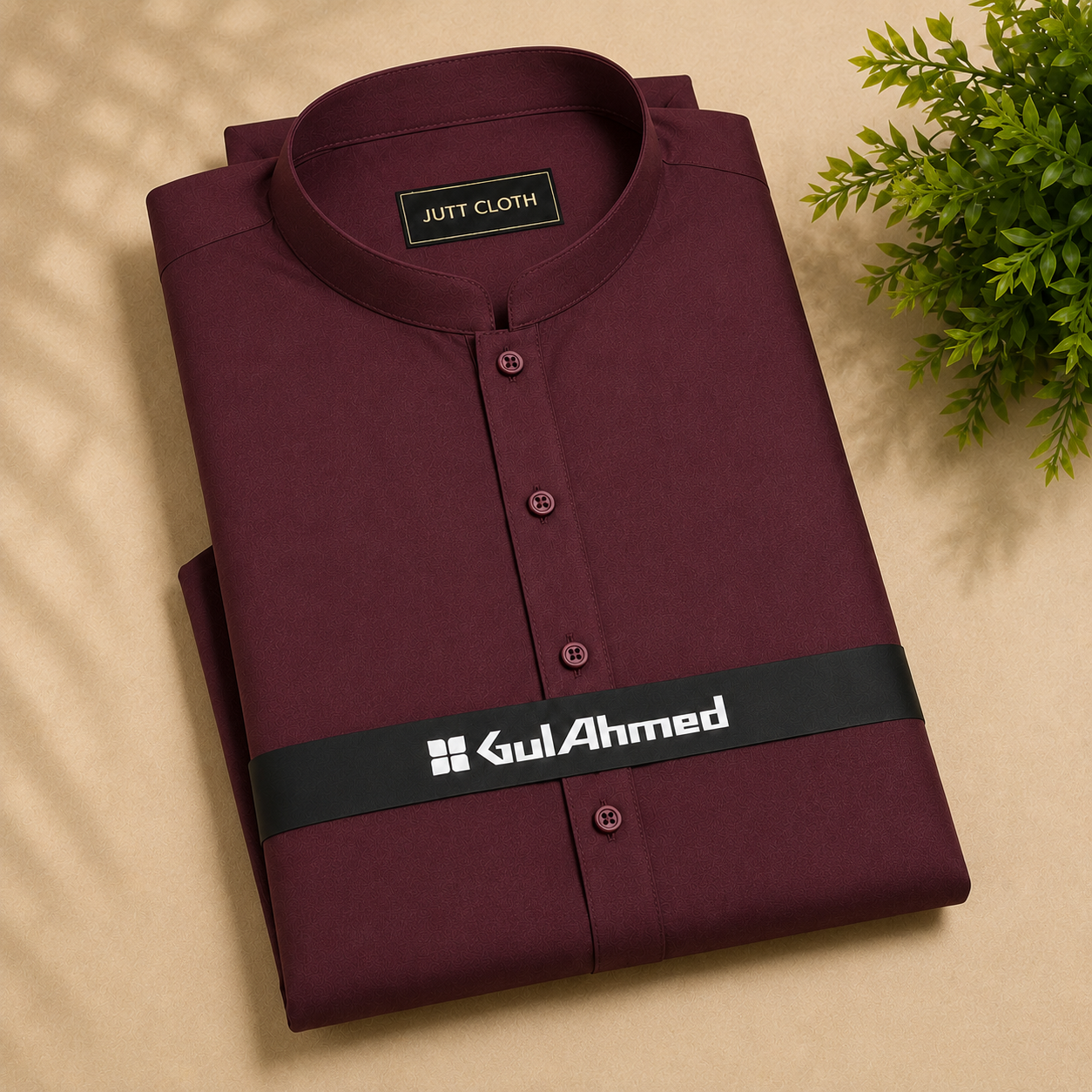 A folded maroon long-sleeve shirt with a mandarin collar and button front, displayed on a beige textured surface with a green plant in the corner. The shirt features a black band across the waist with the white printed brand name 'Gul Ahmed'.