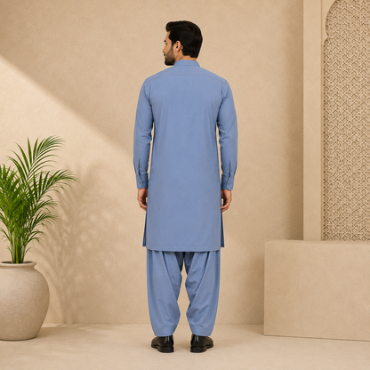 A man viewed from the back wearing a light blue traditional kurta and matching pants, standing in a beige studio setting with a potted plant on the left.