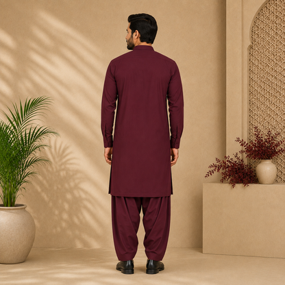 A man viewed from the back wearing a deep maroon long kurta and matching loose trousers, standing in a beige studio set with decorative plants and architectural details.