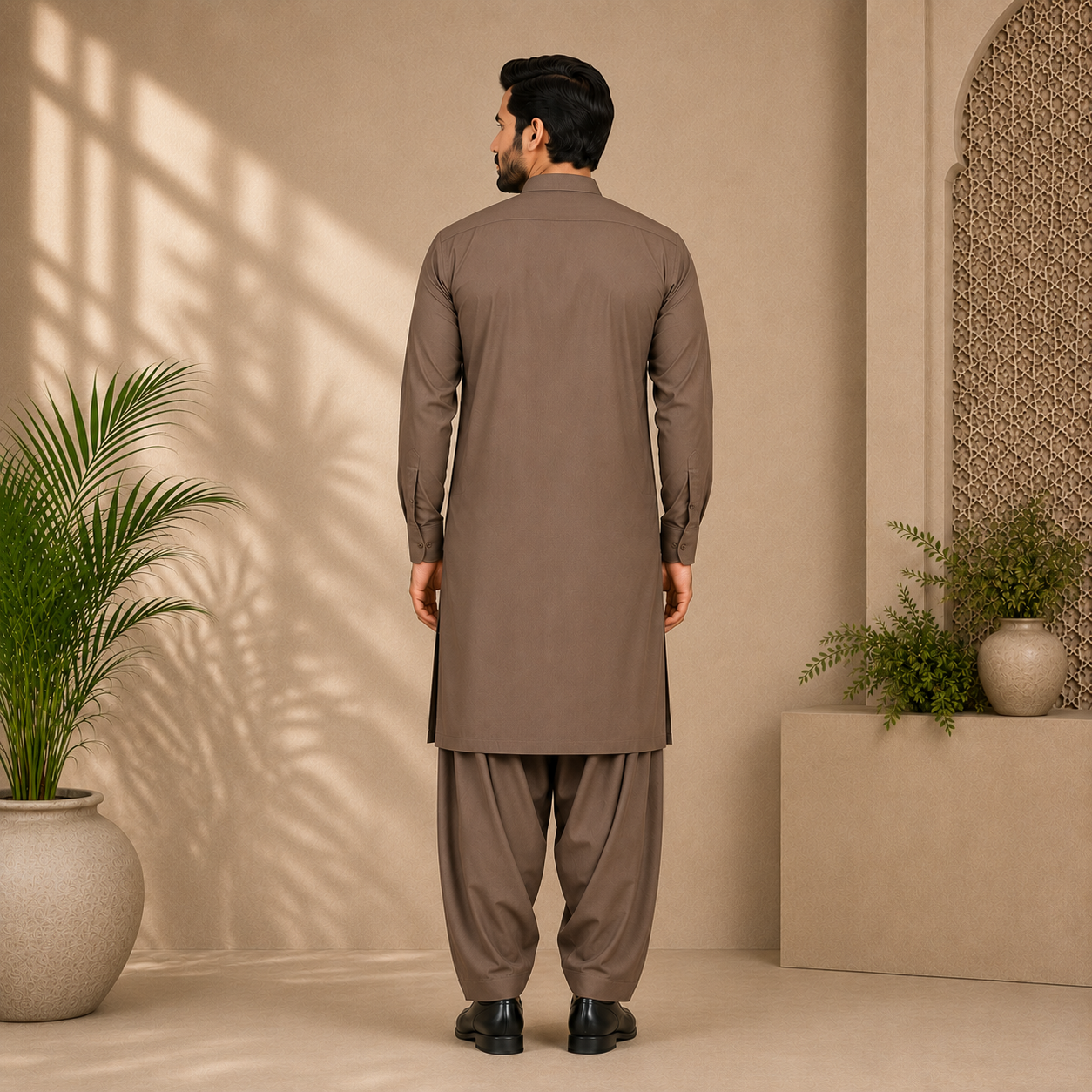 A man viewed from behind wearing a brown long kurta and matching pants in a warm beige interior with plants and soft window shadows.
