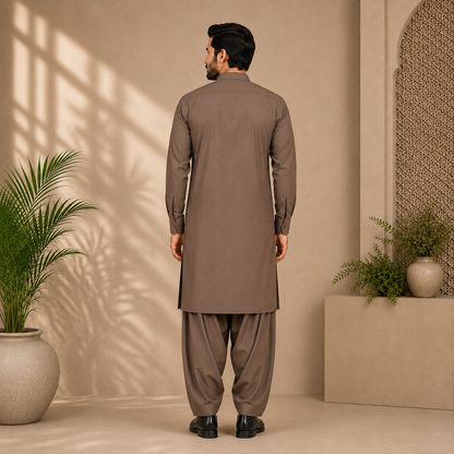 A man viewed from behind wearing a brown long kurta and matching pants in a warm beige interior with plants and soft window shadows.