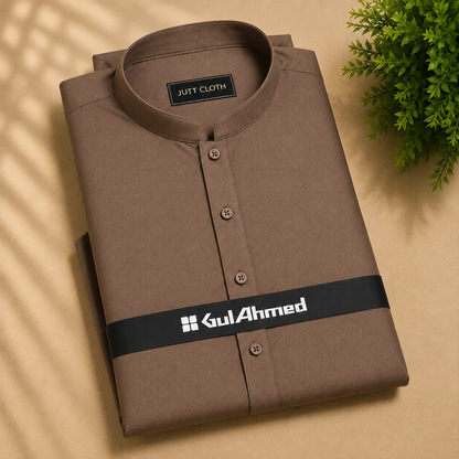 A folded brown kurta shirt with a mandarin collar and button front, displayed on a beige surface with a green plant in the corner. The shirt features a black band around the waist area with the brand name 'Gul Ahmed' printed in white.