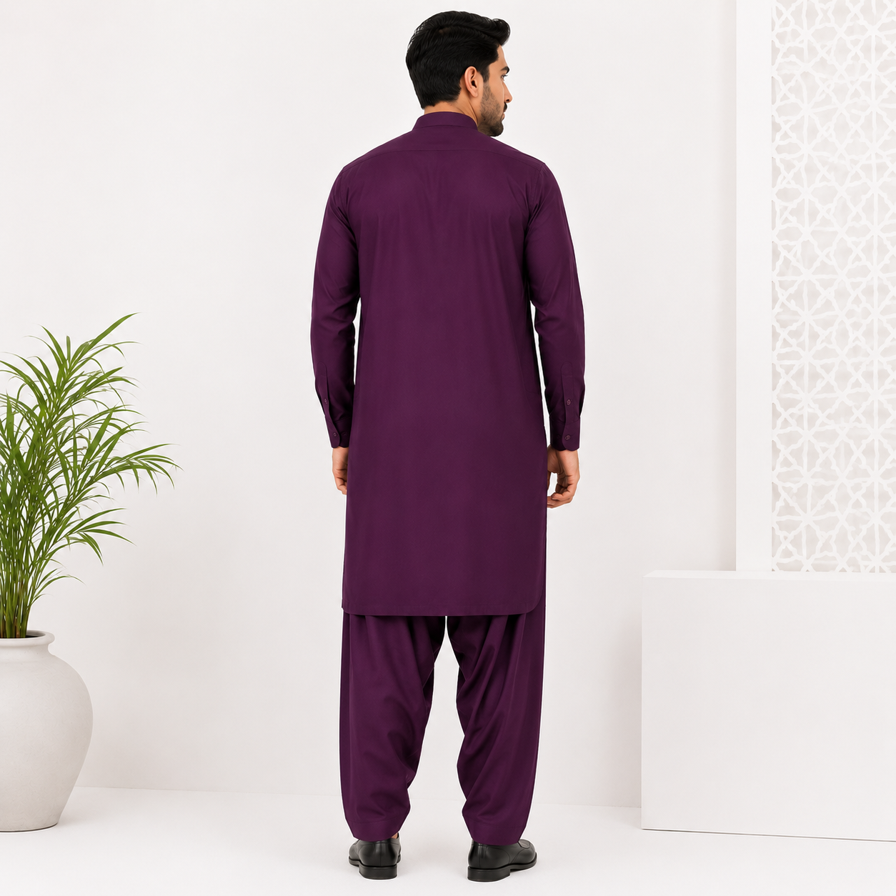 A man viewed from the back wearing a deep purple long kurta and matching pants, standing in a bright white studio with a potted plant on the left and a white geometric panel on the right.