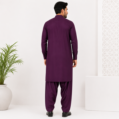 A man viewed from the back wearing a deep purple long kurta and matching pants, standing in a bright white studio with a potted plant on the left and a white geometric panel on the right.