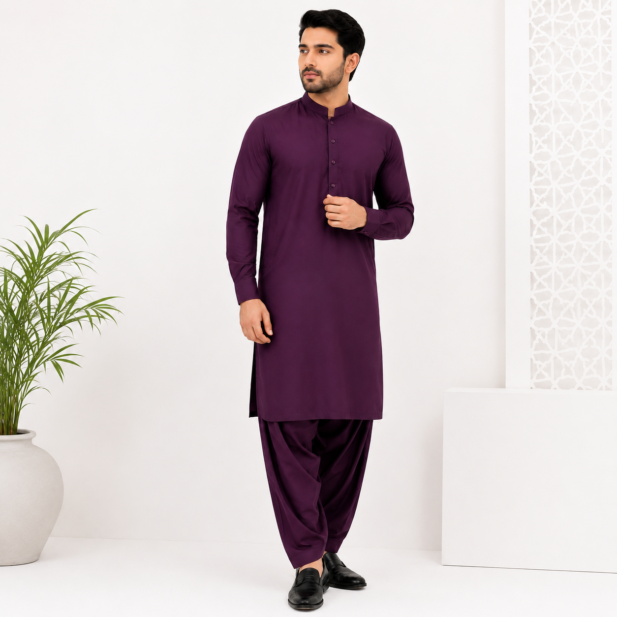 A man modeling a deep purple long-sleeve kurta with matching purple pants, standing in a bright white studio setting with a potted plant on the left.
