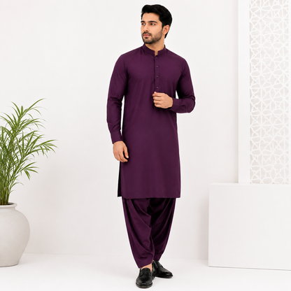 A man modeling a deep purple long-sleeve kurta with matching purple pants, standing in a bright white studio setting with a potted plant on the left.