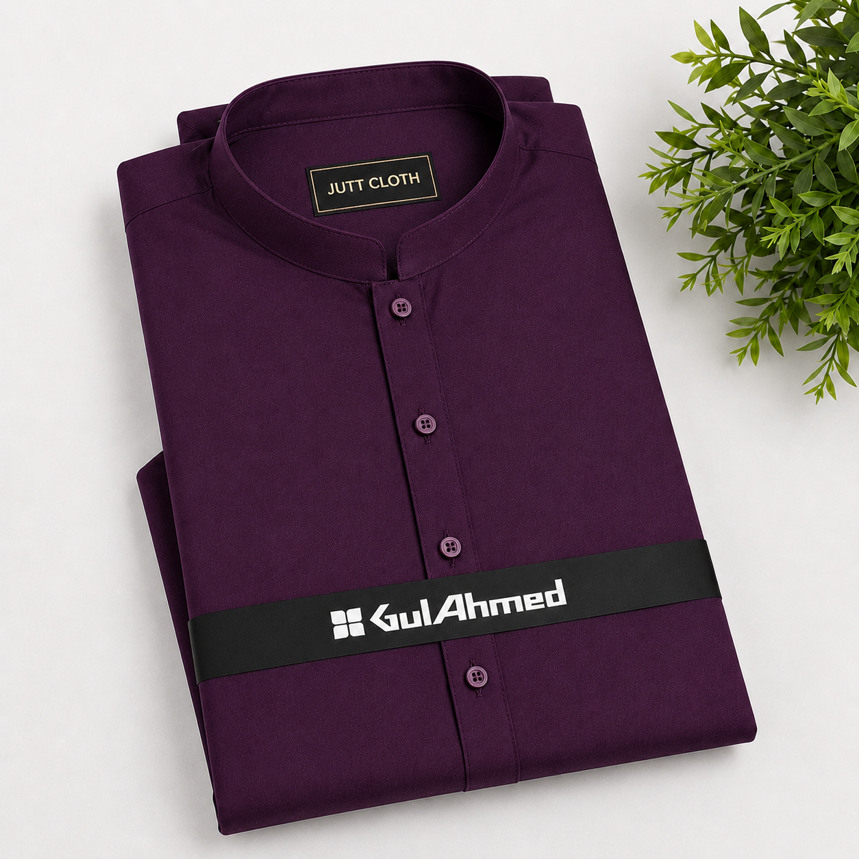 A folded dark purple kurta shirt with a mandarin collar and button front, displayed on a white background with a green plant leaf in the corner. The shirt features a black waistband with the white printed brand name 'GulAhmed'.