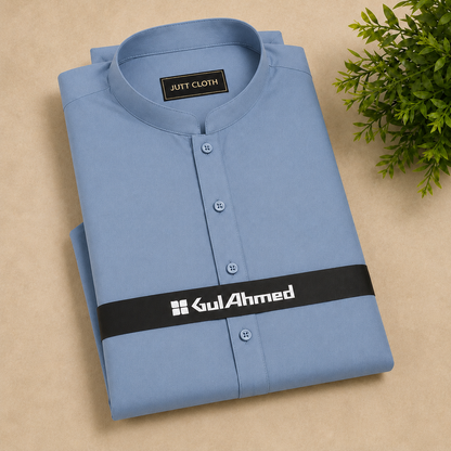 A folded light blue men's shirt with a mandarin collar and button placket, displayed on a beige surface with a small green plant in the corner. The shirt features a black brand band with 'GulAhmed' printed in white.