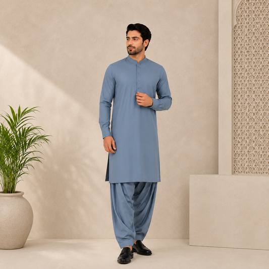 A man modeling a light blue traditional kurta and pant set in a minimalist beige studio setting with a potted plant on the left.