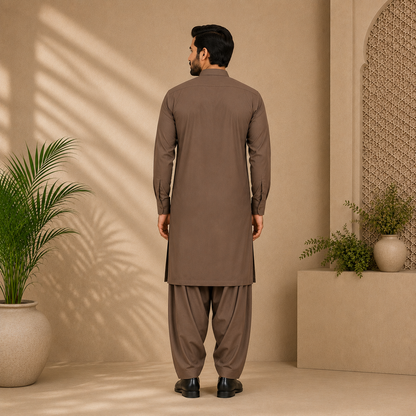 A man stands with his back to the camera wearing a brown long-sleeve kurta and matching wide-leg trousers in a warm beige studio setting with potted plants and soft shadow patterns.